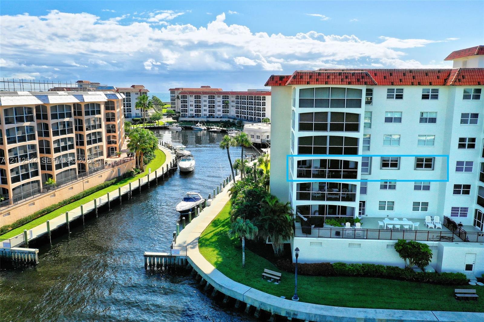 Multi-story waterfront residential building featuring a tile roof, expansive glass-enclosed balconies, and a concrete bulkhead along the canal