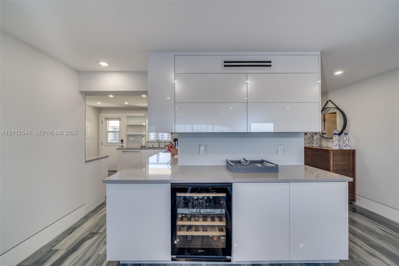 Kitchen island featuring high-gloss white cabinetry, integrated under-counter wine refrigerator, and stone-surface countertop, accented by wood-finish flooring and recessed lighting