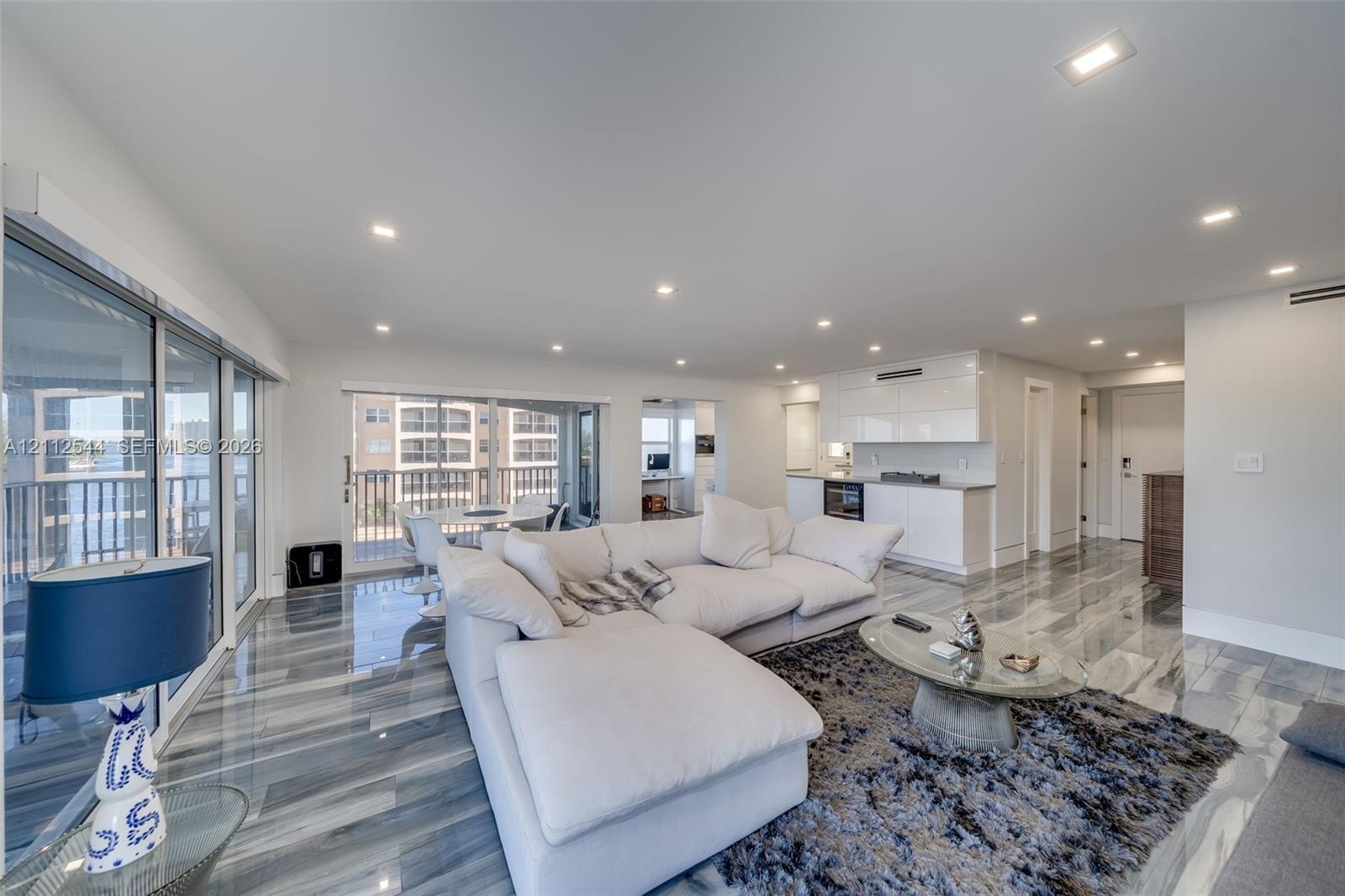 Open-concept living space featuring high-gloss stone flooring, recessed ceiling lighting, sliding glass door access to an exterior balcony, and white cabinetry