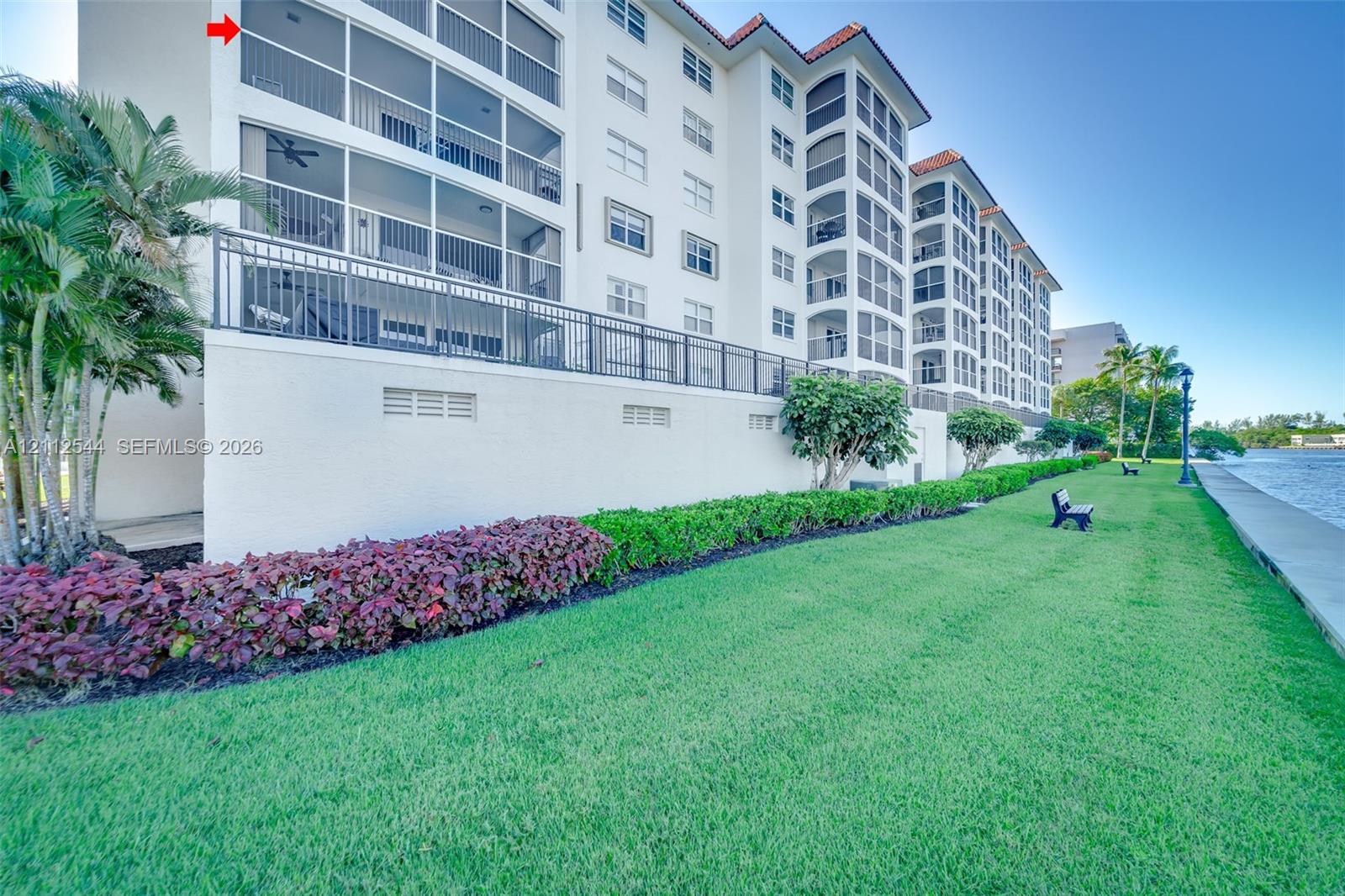 Multi-story building exterior featuring screened-in balconies, a stucco facade, red tile roofing, and direct waterfront grounds with manicured landscaping