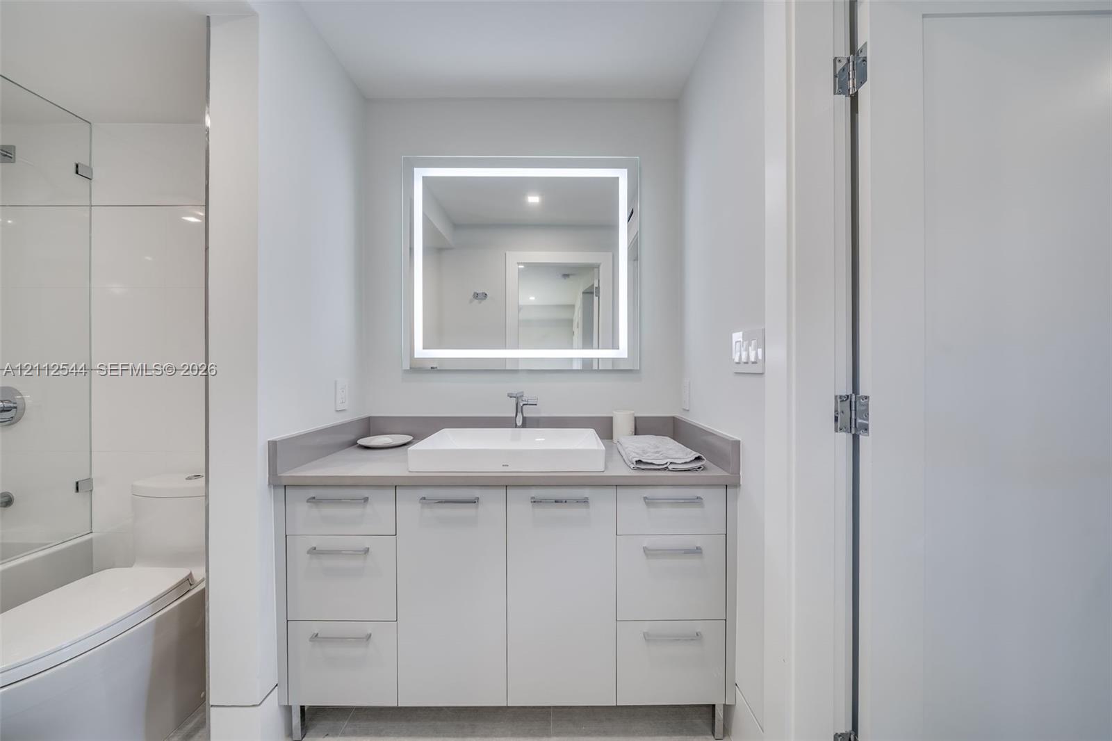 Bathroom vanity featuring flat-panel cabinetry, a rectangular vessel sink, solid-surface countertop, and an integrated LED-backlit mirror