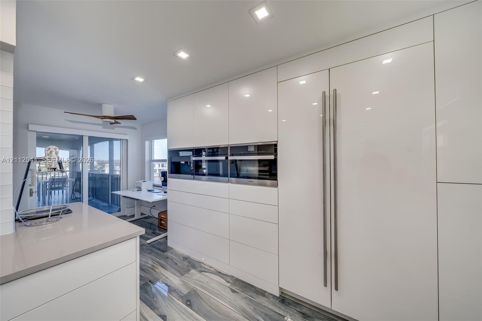 Sleek, high-gloss white cabinetry featuring integrated, full-height refrigerator and flush-mount wall ovens, quartz countertops, recessed ceiling lighting, and polished stone-finish flooring
