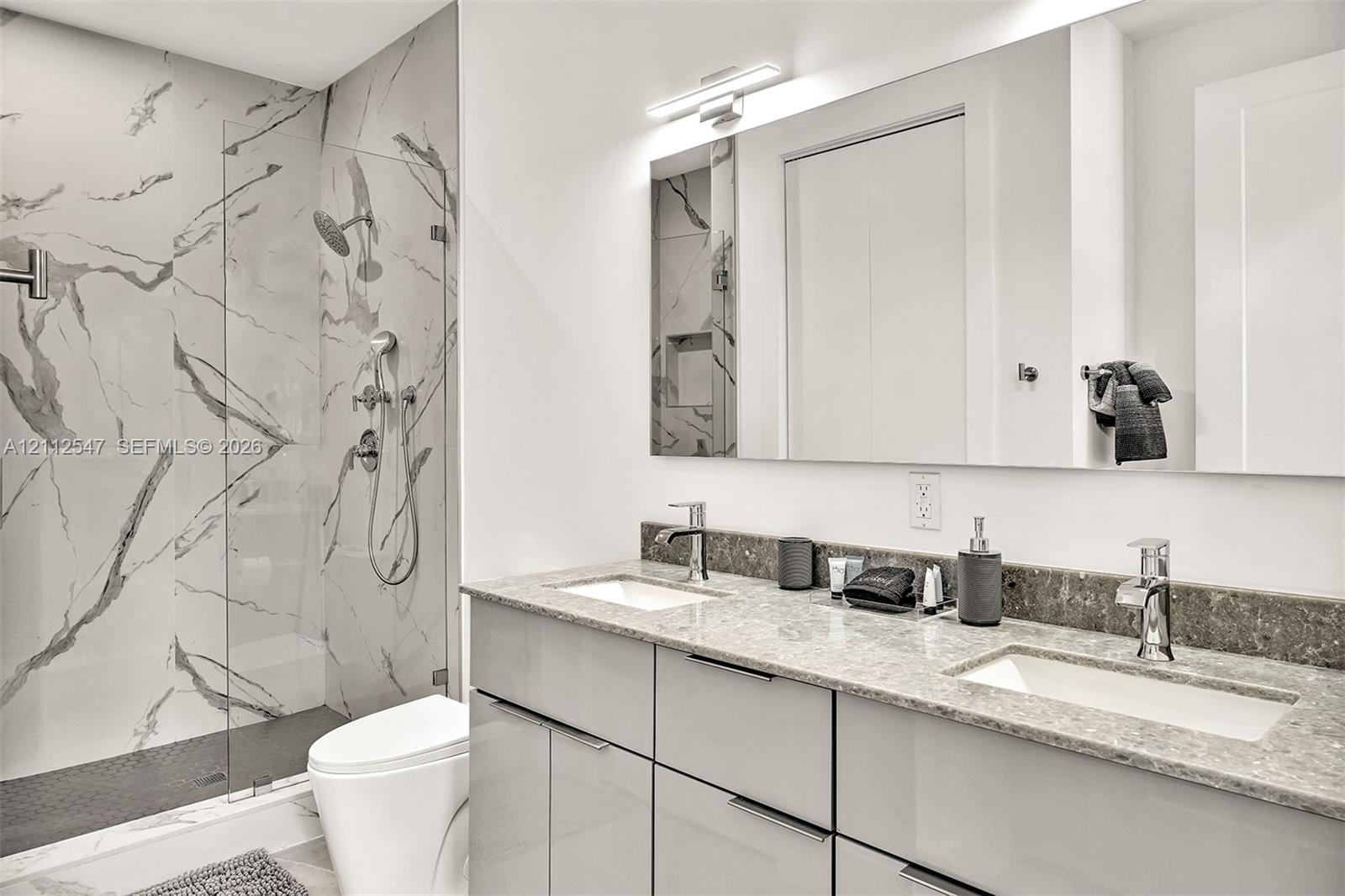 Bathroom featuring a dual-sink vanity with stone countertop, walk-in shower with floor-to-ceiling tile surround, glass partition, and wall-mounted light fixture