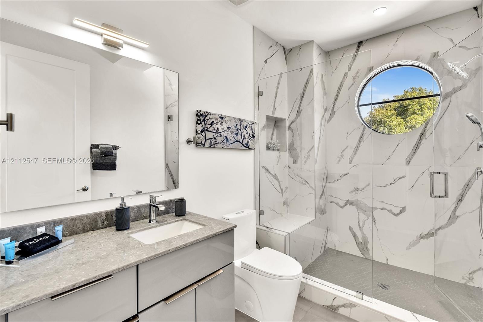 Bathroom featuring a sleek vanity with a stone countertop, an integrated rectangular sink, wall-to-wall mirror, walk-in shower with floor-to-ceiling stone-patterned wall tile, and a decorative circular window