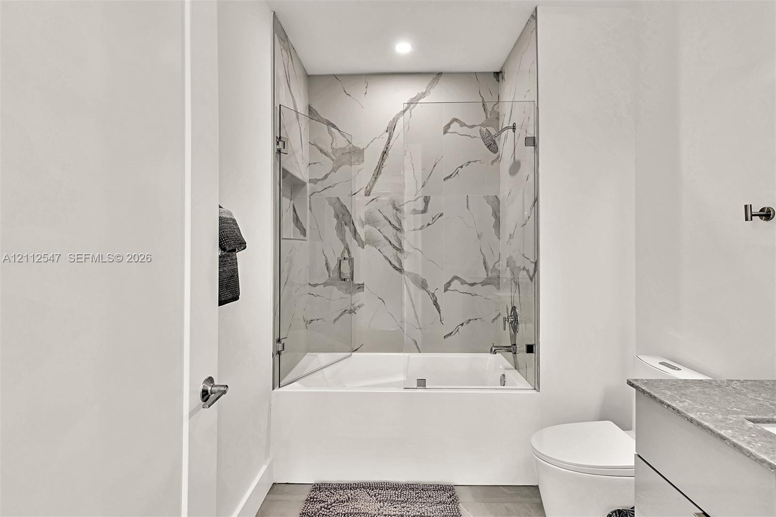 Full-height shower surround with veined tile, frameless glass enclosure, recessed shower niche, deep soaking tub, and stone-finish vanity countertop