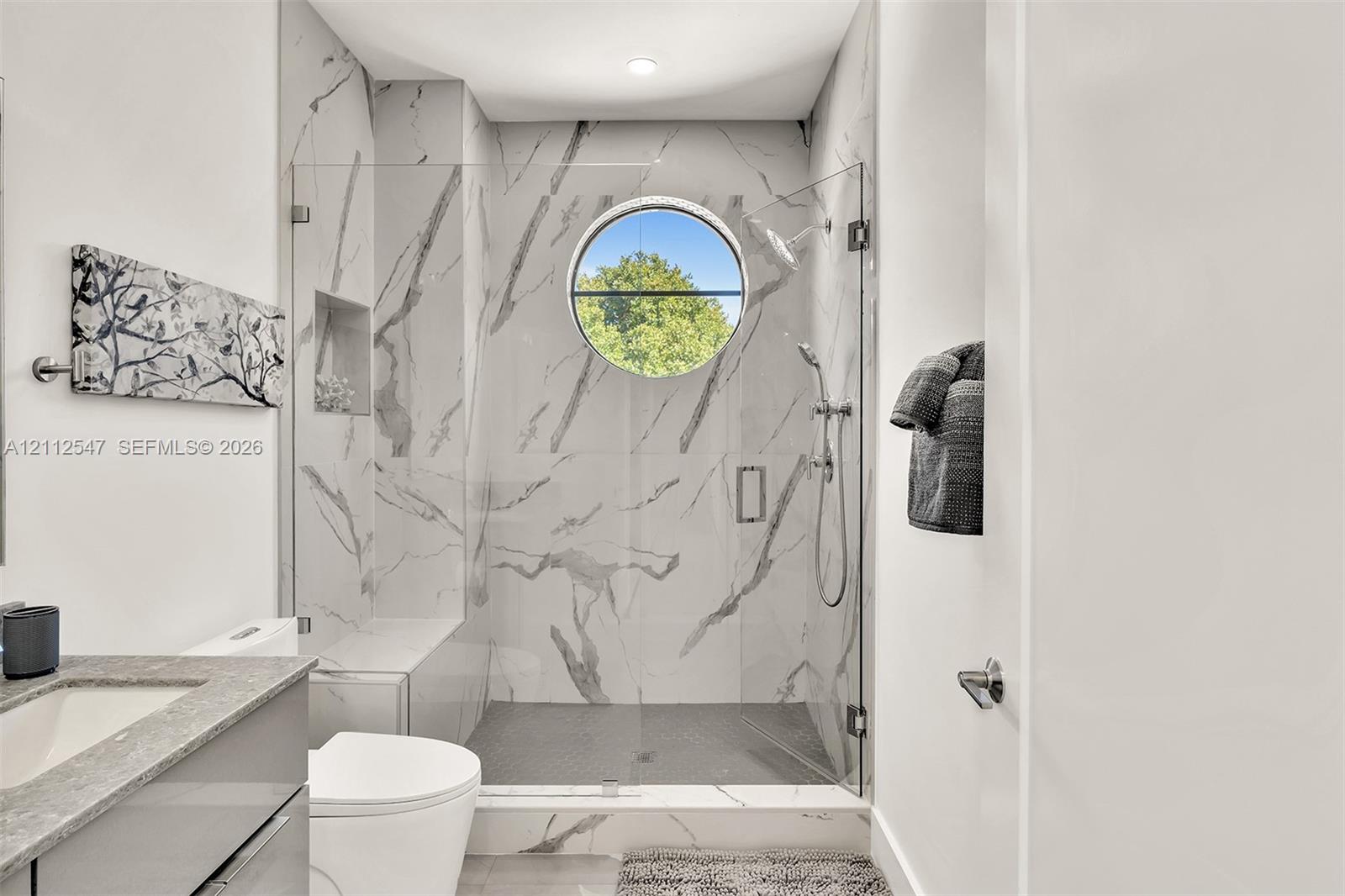 Walk-in shower featuring large-format patterned tile, glass enclosure, built-in bench, circular window, and handheld shower fixture