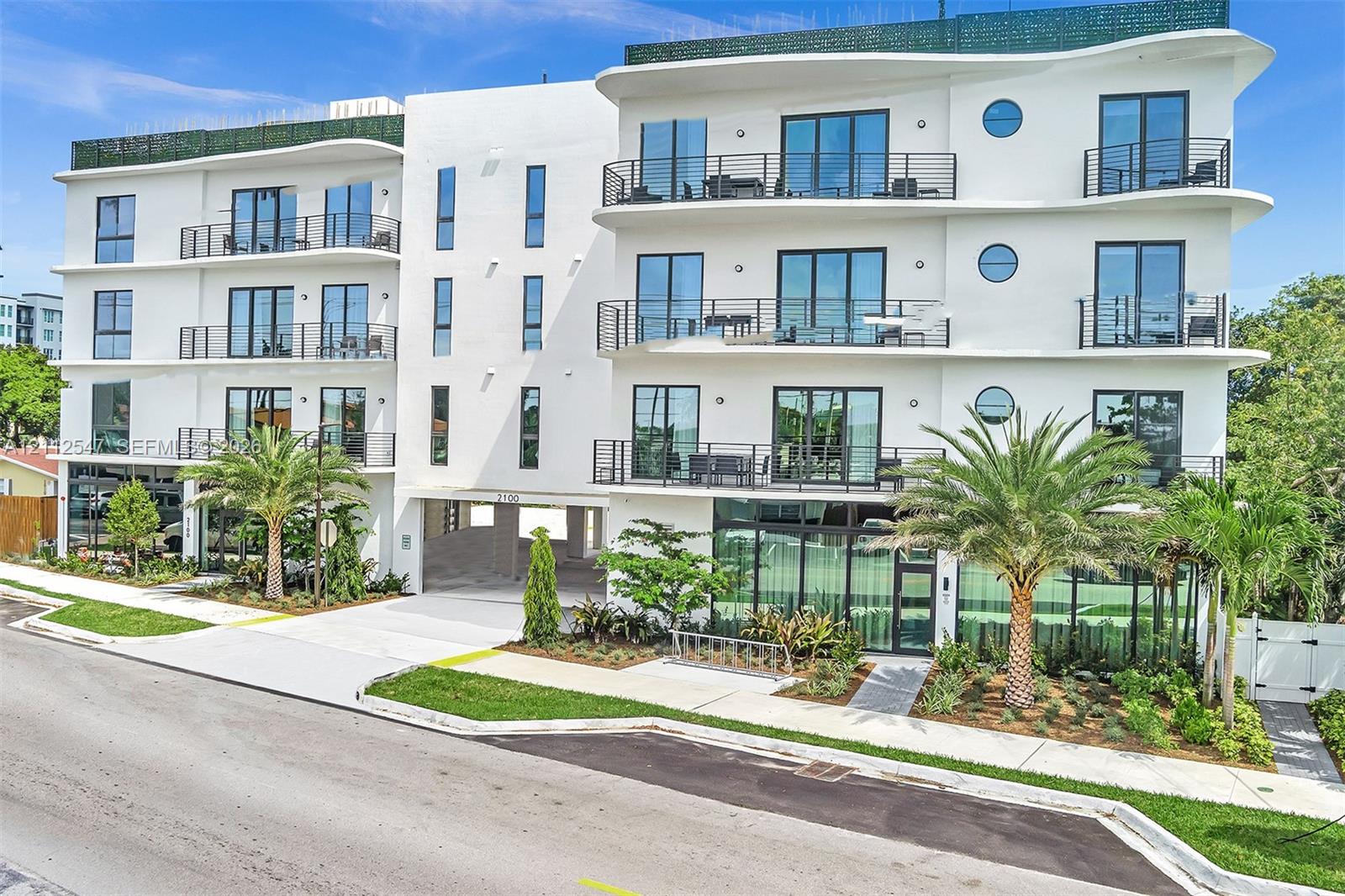 Contemporary mid-rise exterior featuring smooth white stucco finish, wraparound balconies with metal railing, floor-to-ceiling glass windows, circular architectural accents, and manicured landscaping