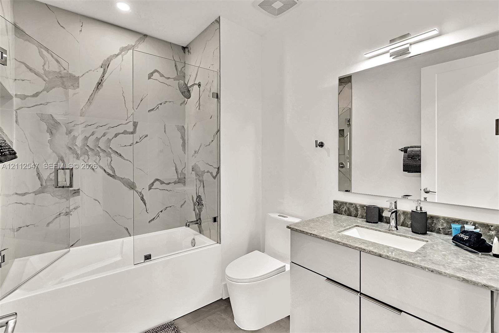 Bathroom featuring a tub-shower combination with floor-to-ceiling large-format tile surround, a glass shower panel, a floating vanity with a stone countertop and rectangular undermount sink, and an expansive wall-mounted mirror