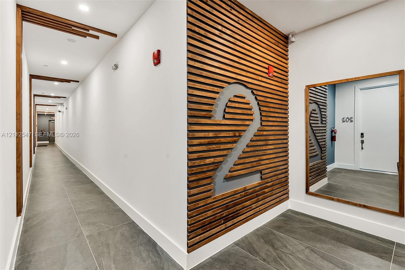 Hallway featuring large-format tile flooring, horizontal wood-slat accent wall, integrated recessed ceiling lighting, and wood-framed mirror