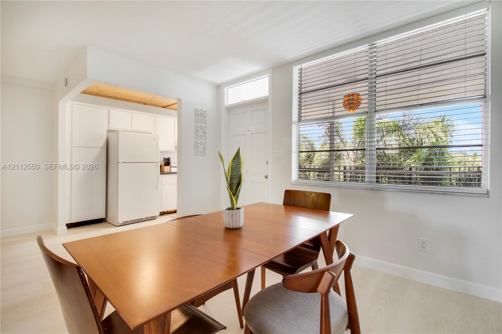 Bright room featuring wood-finish flooring, a large window with horizontal blinds, and access to a kitchen with white cabinetry