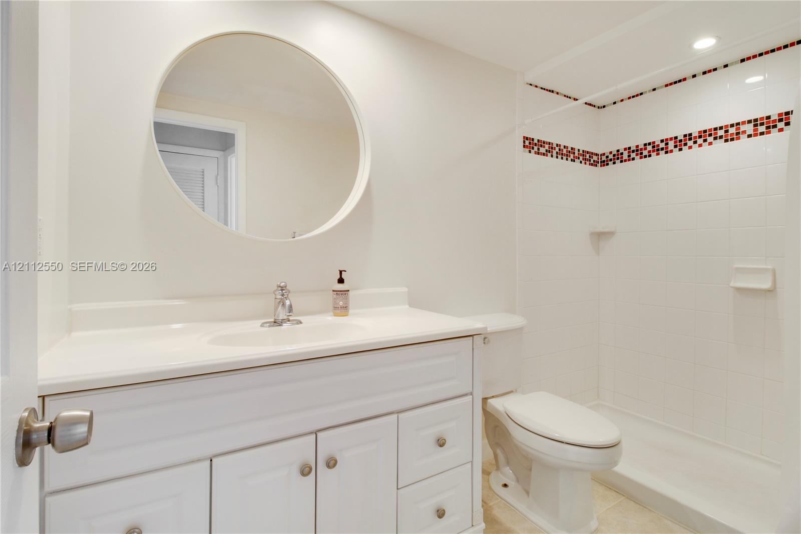 White vanity featuring a solid-surface countertop with an integrated basin, circular wall-mounted mirror, and a tile shower surround with decorative mosaic accents