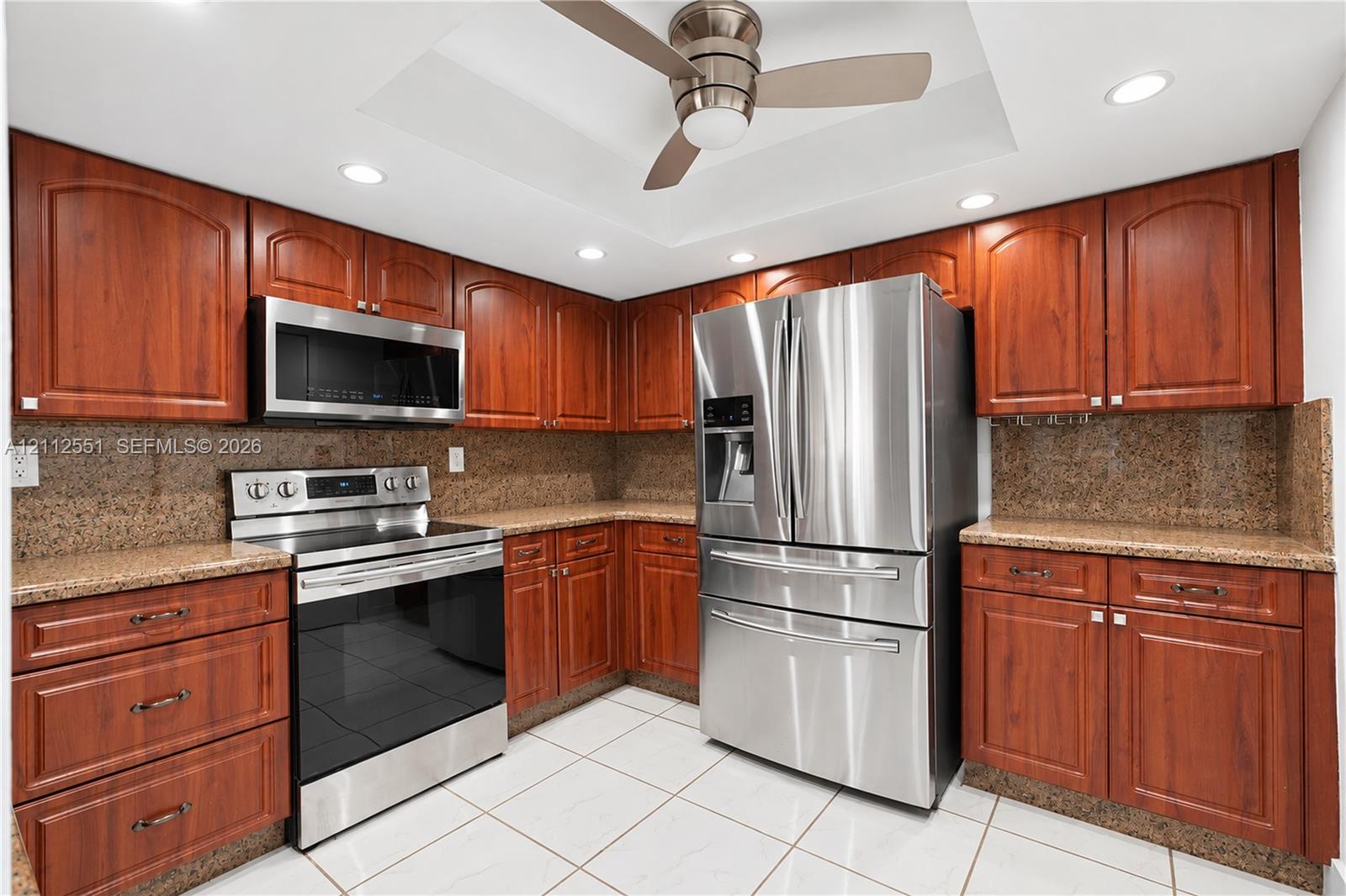 Kitchen featuring cherry-finish cabinetry, stone countertops, stainless steel appliances, a recessed ceiling with a central fan, and tile flooring Kitchen featuring cherry-finish cabinetry, stone countertops, stainless steel appliances, a recessed ceiling with a central fan, and tile flooring