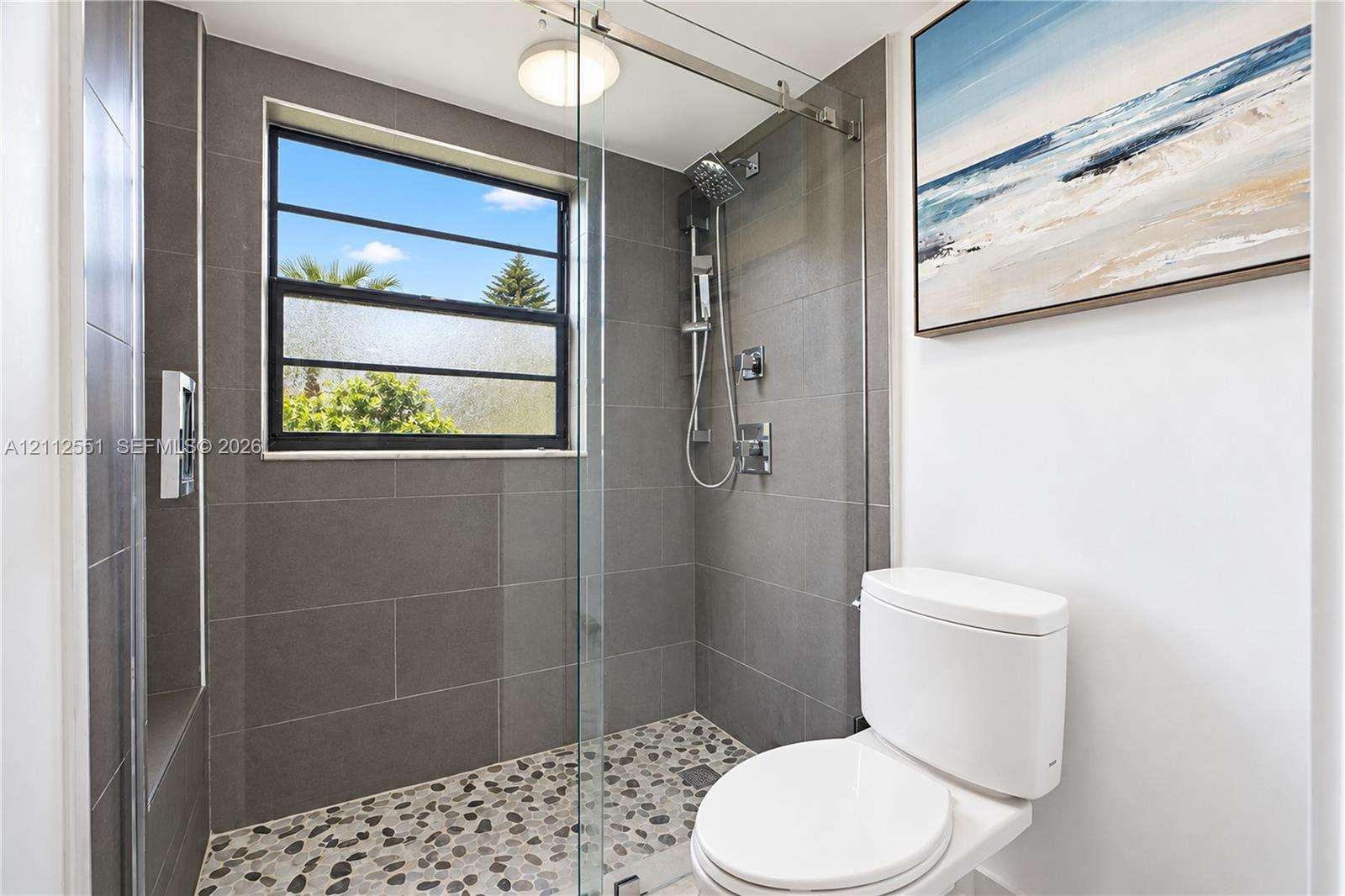 Walk-in shower featuring large-format grey wall tile, a pebble-stone floor, glass panel enclosure, and a chrome rainfall shower head