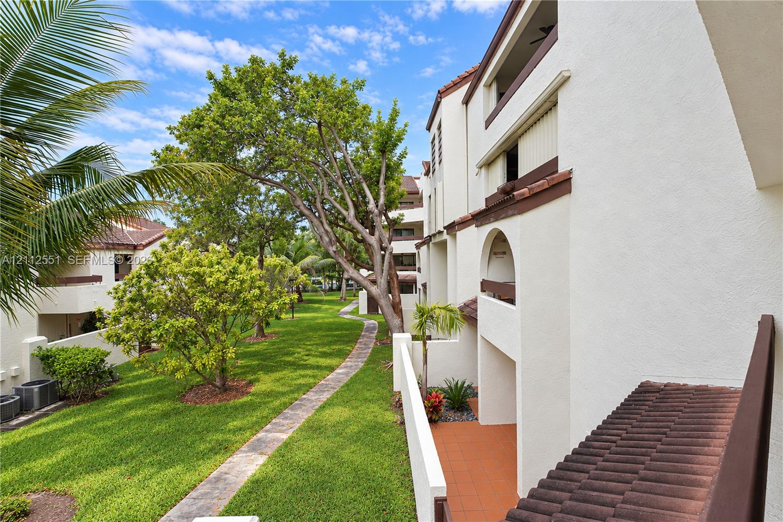 Stucco exterior featuring arched architectural detailing, terracotta-tiled roof accents, private tiled balconies, and a paved walkway through a manicured garden setting