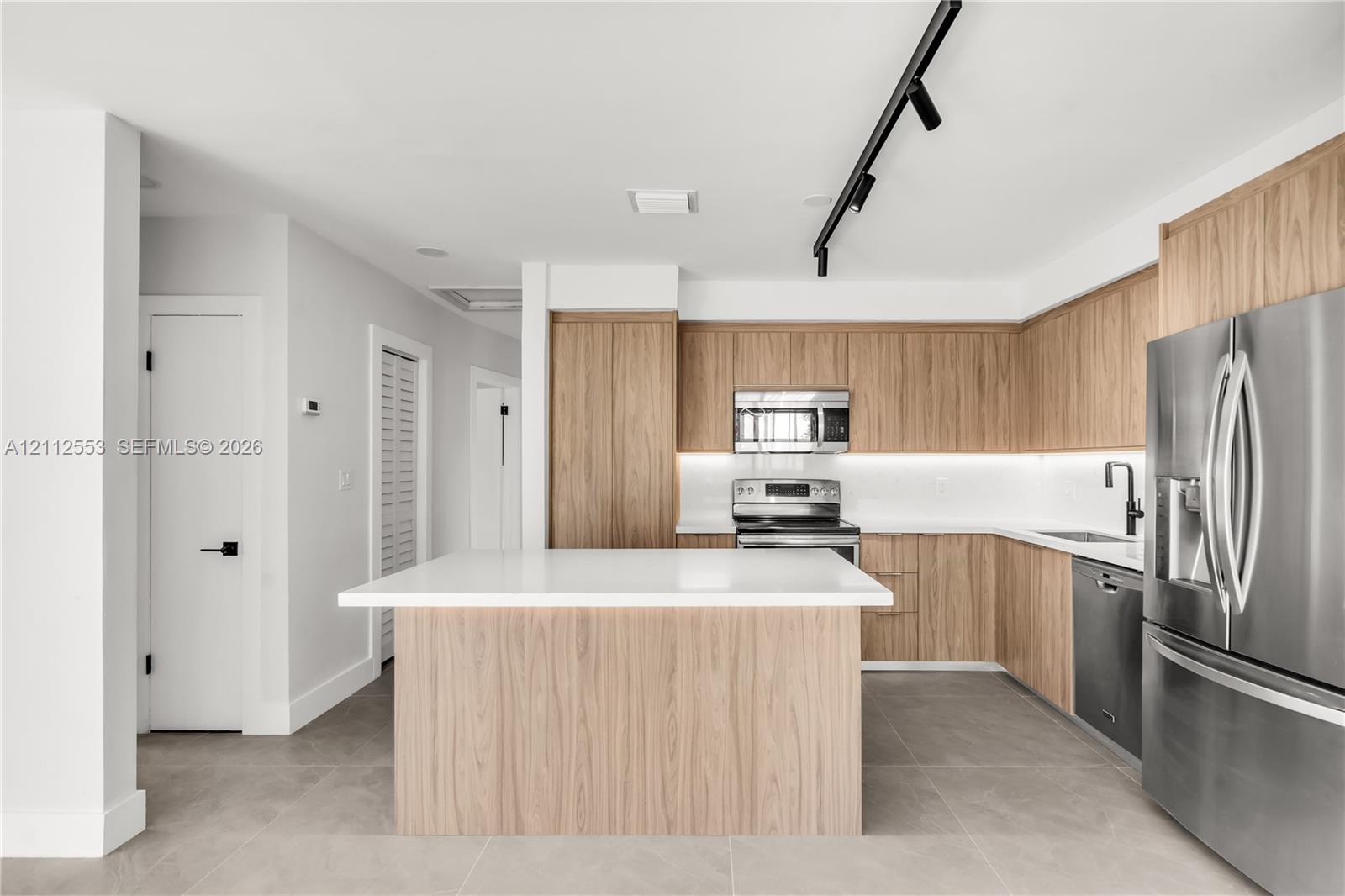 Kitchen featuring wood-finish cabinetry, a central island with a solid surface countertop, stainless steel appliances, neutral floor tiling, and black track lighting