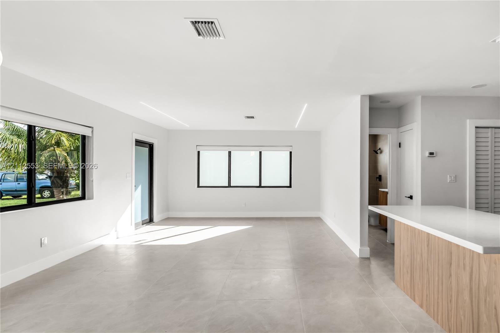 Open interior featuring large-format tile flooring, recessed linear lighting, expansive windows with black frames, and a kitchen island with a light-toned wood-finish base and quartz countertop