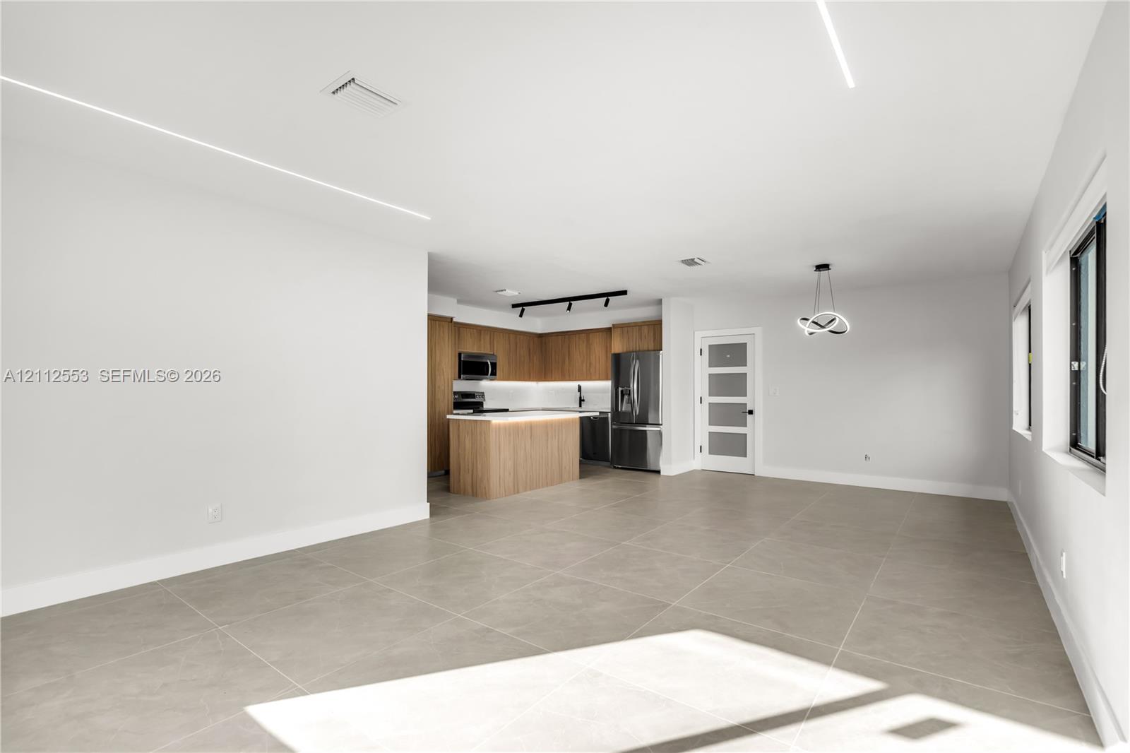 Open-concept interior featuring large-format tile flooring, recessed linear lighting, kitchen with wood-finish cabinetry, center island, and a multi-pane frosted glass door
