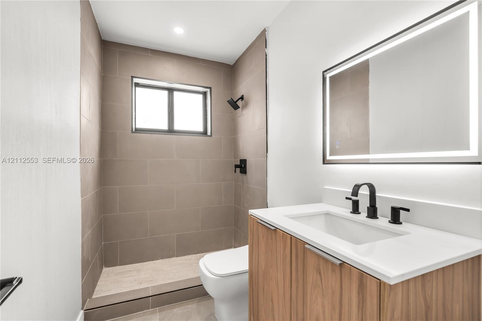 Modern vanity with wood-finish cabinetry, white countertop, rectangular undermount sink, and matte black widespread faucet, complemented by an LED-backlit mirror and a walk-in shower featuring large-format tile surround and matte black fixtures