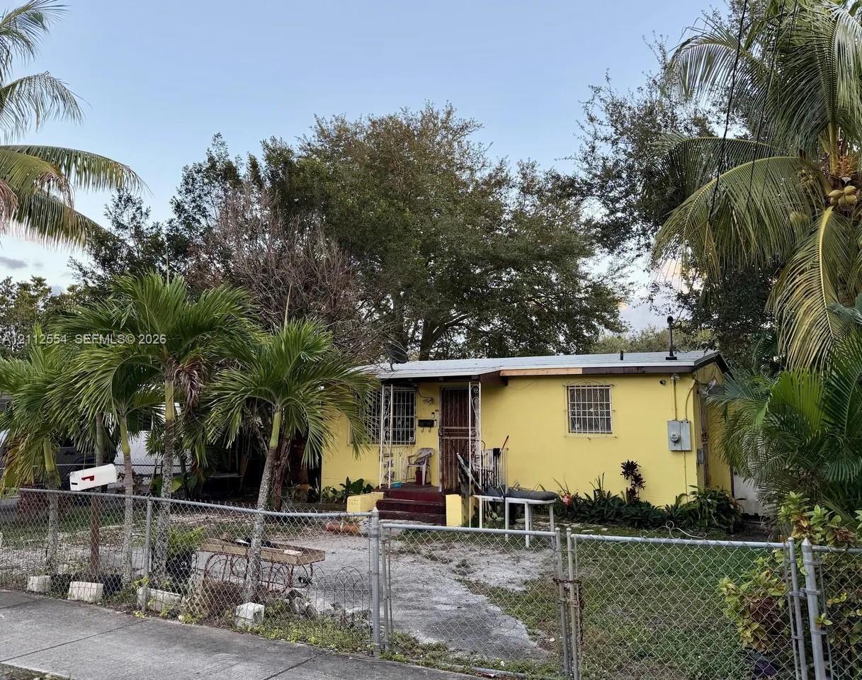 Single-story residence featuring a painted stucco exterior, metal-framed window guards, covered front entryway, and mature palm landscaping situated within a chain-link fenced perimeter