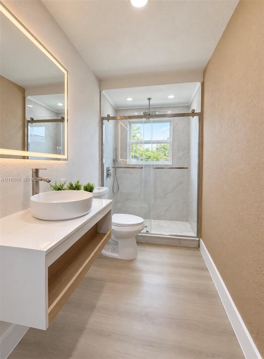 Floating vanity with an integrated vessel sink, illuminated wall mirror, glass-enclosed shower featuring large-format tile surround, and light-toned wood-finish flooring