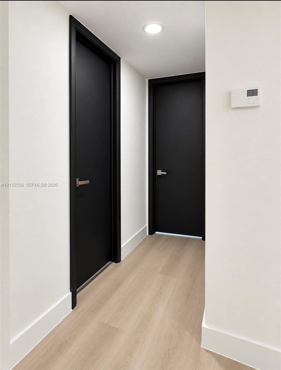 Hallway featuring light-toned wood-finish flooring, solid matte black doors with minimalist metal lever handles, recessed ceiling lighting, and smooth white walls