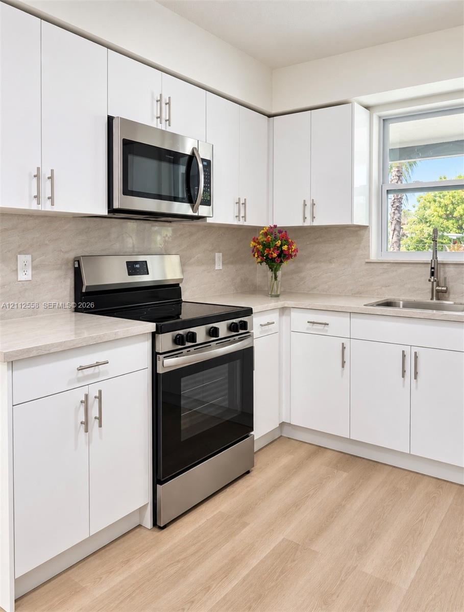 Kitchen featuring white shaker-style cabinetry, stainless steel appliances, neutral stone-patterned backsplash, light-toned countertop, and wood-finish flooring Kitchen featuring white shaker-style cabinetry, stainless steel appliances, neutral stone-patterned backsplash, light-toned countertop, and wood-finish flooring