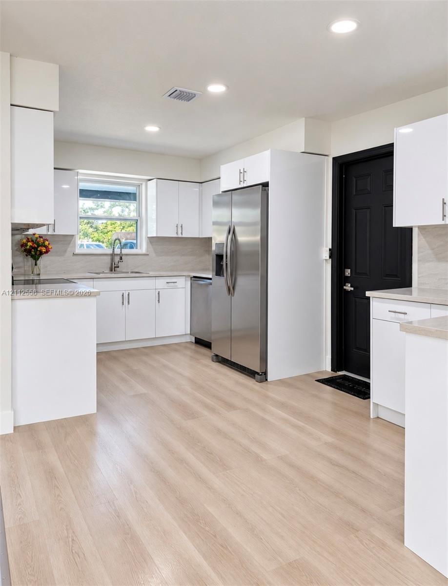 Kitchen featuring high-gloss white cabinetry, light-toned wood-finish flooring, stainless steel appliances, neutral stone-finish countertops, and recessed lighting Kitchen featuring high-gloss white cabinetry, light-toned wood-finish flooring, stainless steel appliances, neutral stone-finish countertops, and recessed lighting
