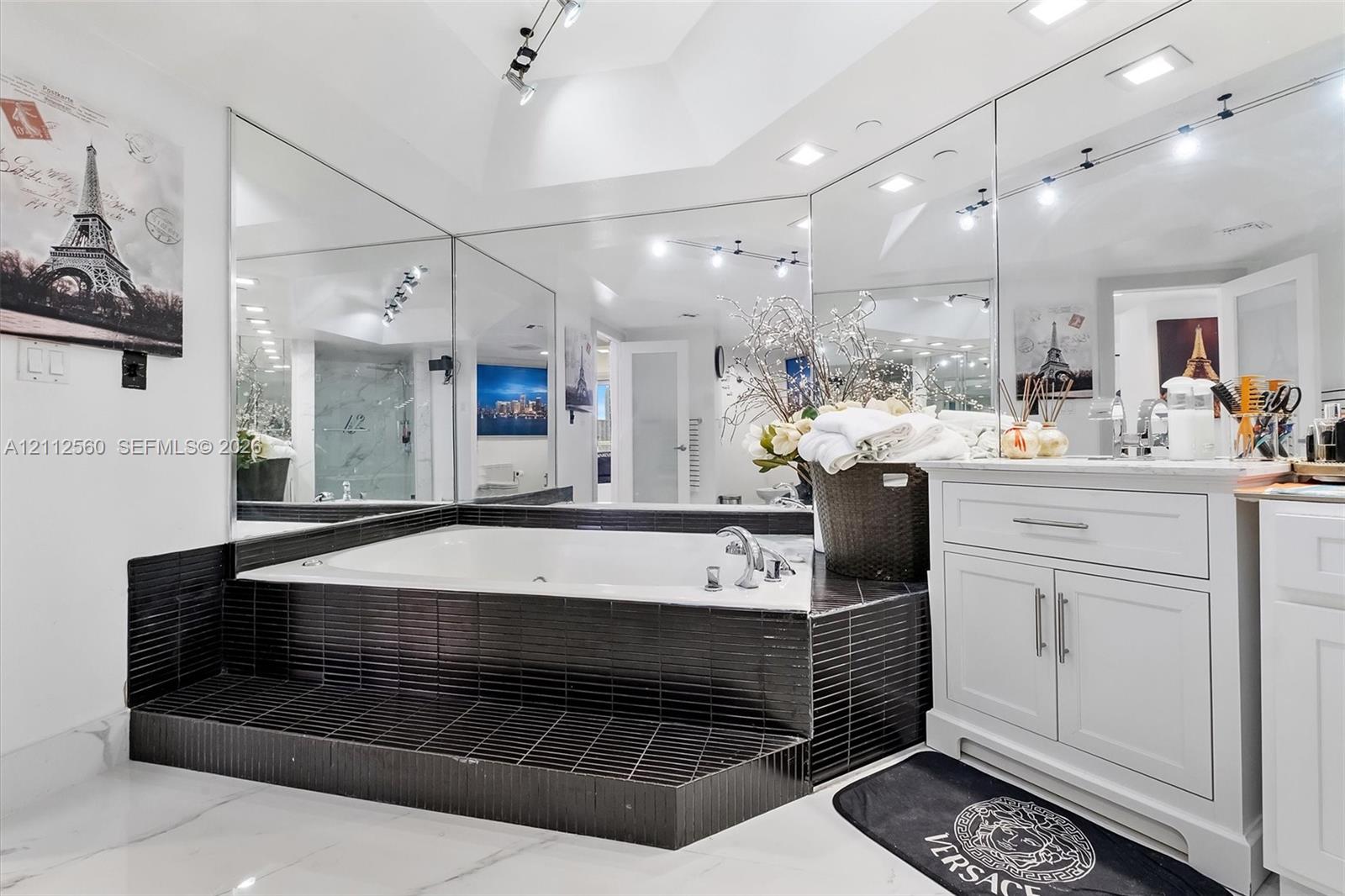Sunken jetted tub finished with black linear tile, white cabinetry with silver hardware, full-width wall mirrors, recessed lighting, and large-format flooring