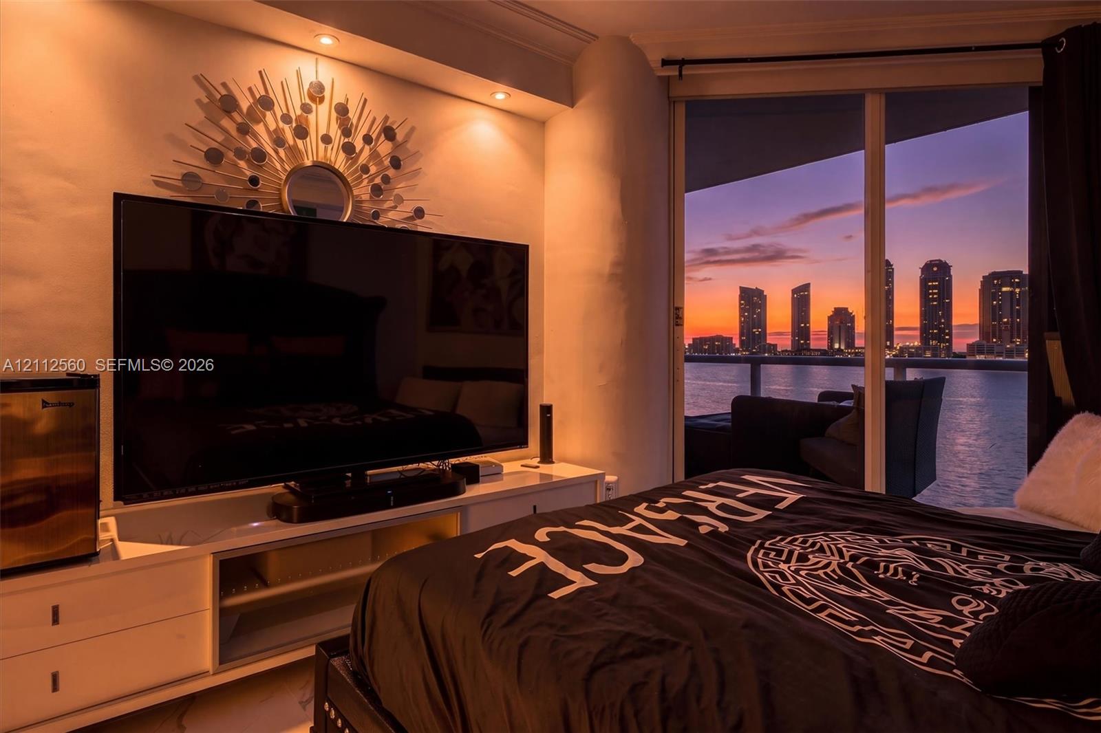 Floor-to-ceiling glass sliding doors, built-in recessed ceiling lighting, and private balcony with metal railing, set against a waterfront city skyline backdrop