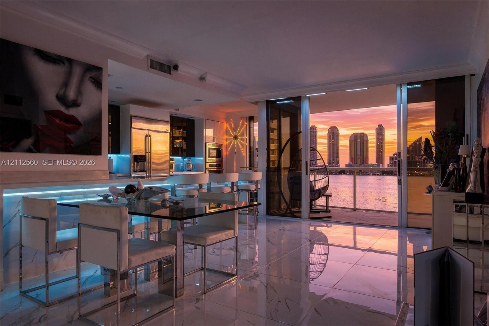 Large-format polished tile flooring, integrated linear LED under-cabinet lighting, stainless steel built-in appliances, and expansive sliding glass doors opening to a balcony with waterfront and skyline scenery