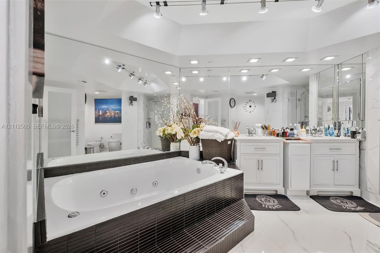 Jetted soaking tub with black tile surround, double vanity with white cabinetry, expansive wall-to-wall mirrors, recessed lighting, and large-format flooring