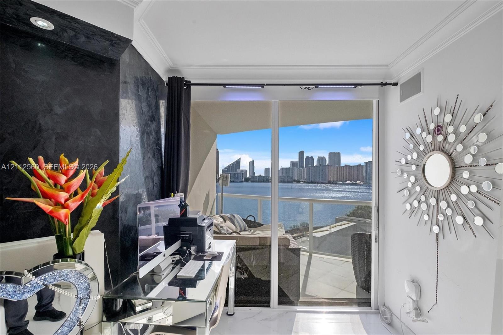 Floor-to-ceiling glass sliding doors opening to a balcony with water and skyline scenery, featuring a textured black feature wall, recessed lighting, and crown molding