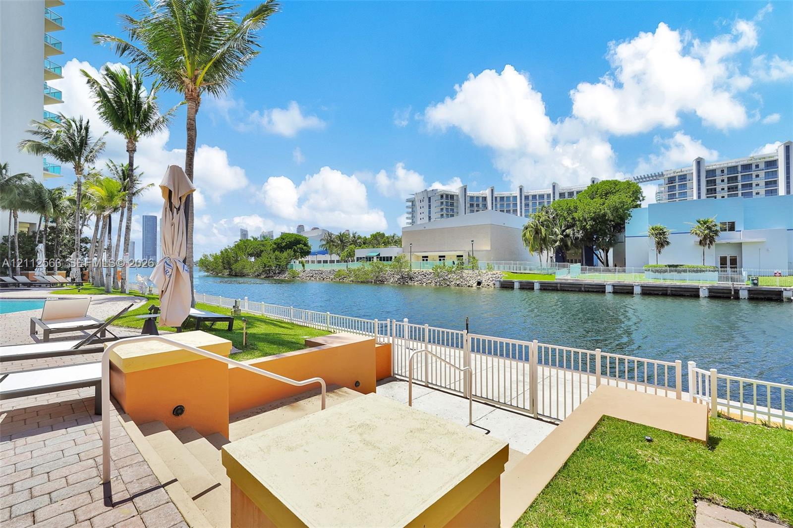Waterfront patio area featuring paved stone decking, built-in tiered retaining walls, a metal safety railing, and mature palm trees