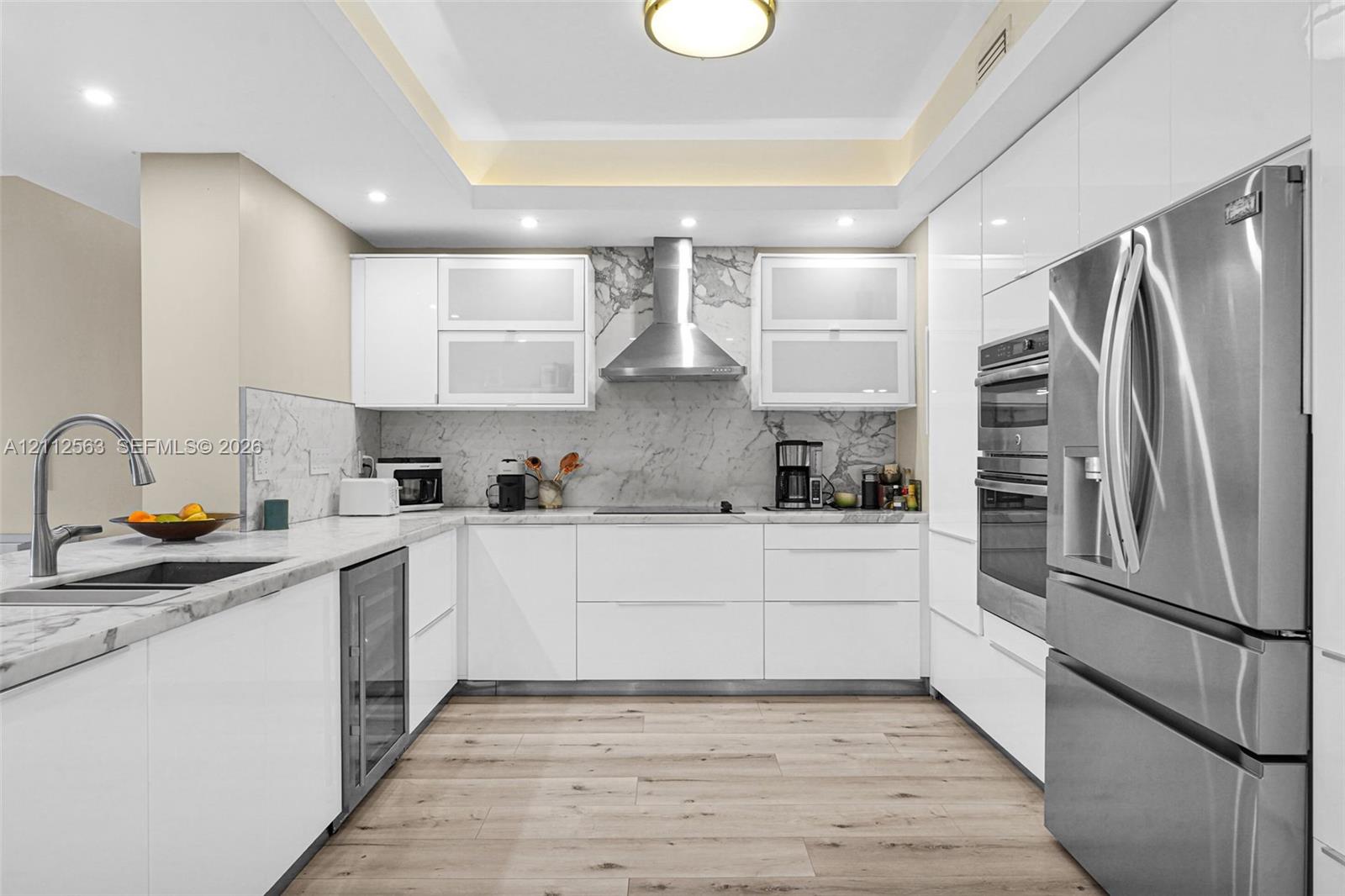 Contemporary kitchen featuring high-gloss white cabinetry, stone slab backsplash, stainless steel appliances, built-in dual ovens, and wood-finish flooring