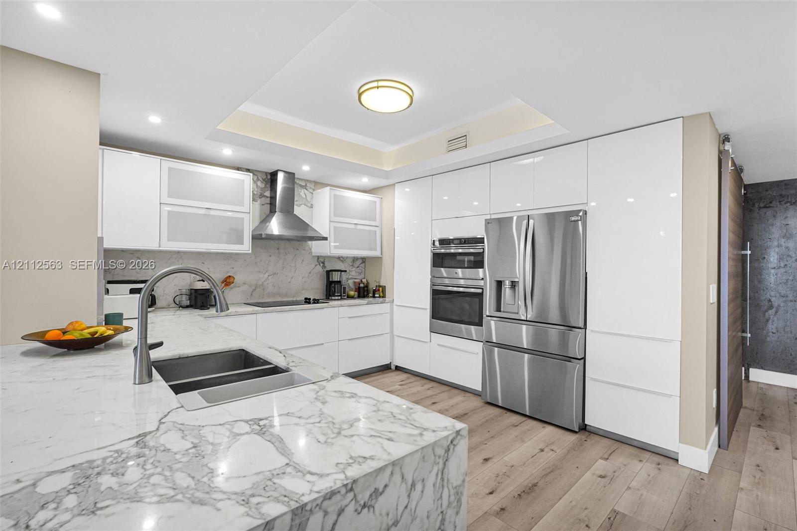 Modern kitchen featuring floor-to-ceiling high-gloss cabinetry, stone countertops, stainless steel appliance suite, wood-finish flooring, and a recessed ceiling design