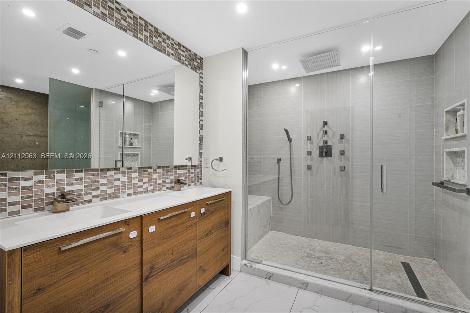 Double vanity featuring wood-finish cabinetry, solid-surface countertops, and a full-width mirror with mosaic tile accents