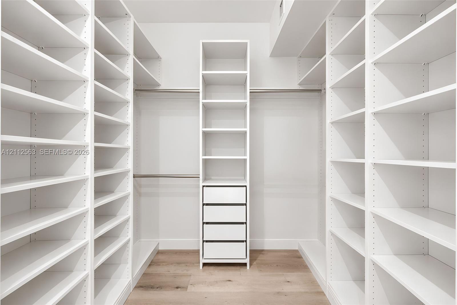 Walk-in closet featuring floor-to-ceiling white built-in shelving, integrated four-drawer stack, metal hanging rods, and wood-finish flooring