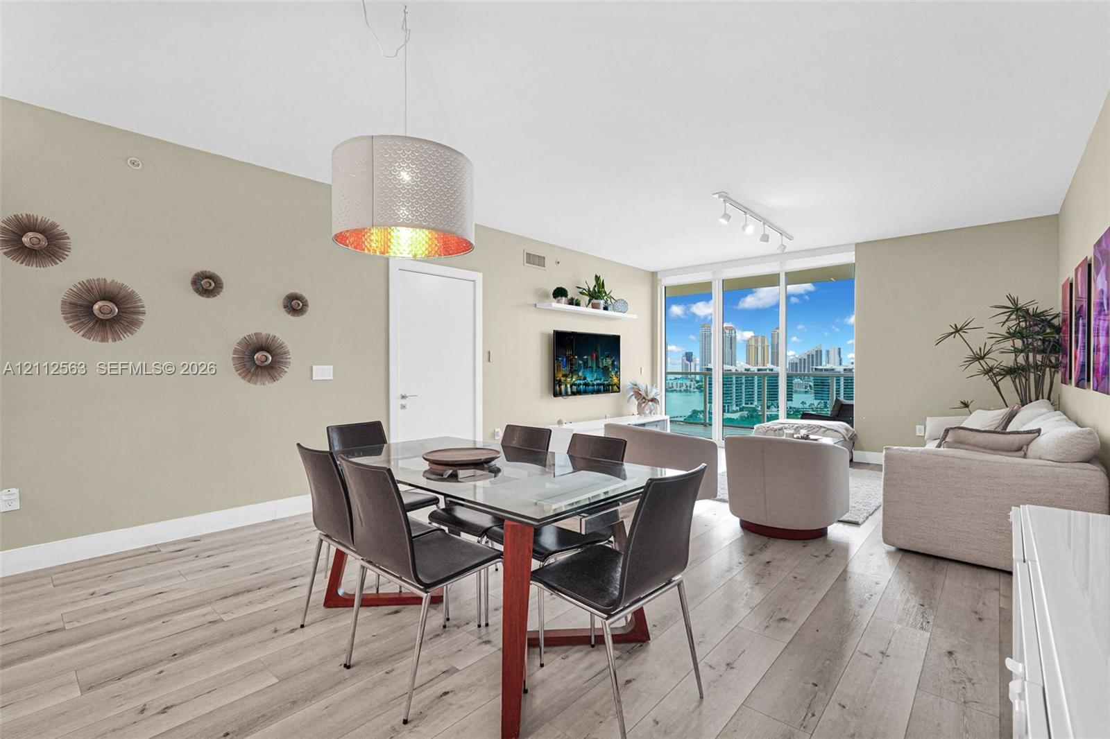 Open-concept living space featuring wood-finish flooring, floor-to-ceiling glass doors, cylindrical pendant lighting, a floating wall-mounted shelf, and a glass balcony railing