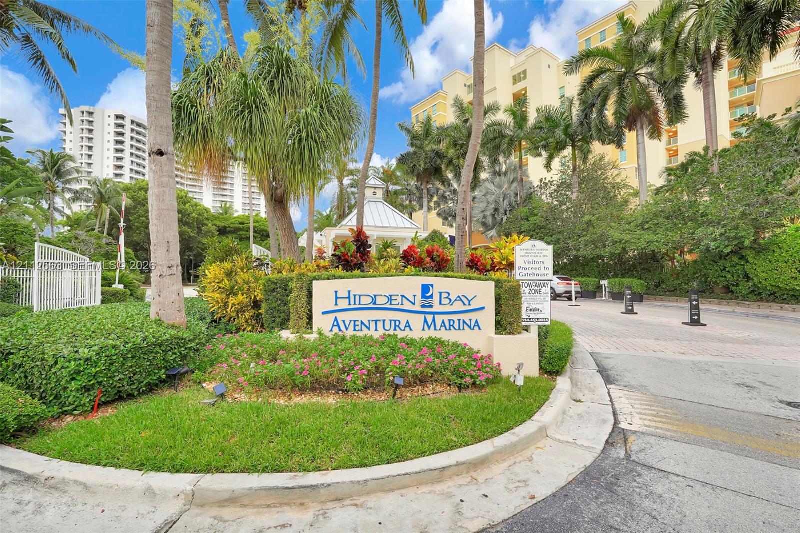 Gated entry featuring stone-finish monument signage, manicured tropical landscaping, and mature palm trees