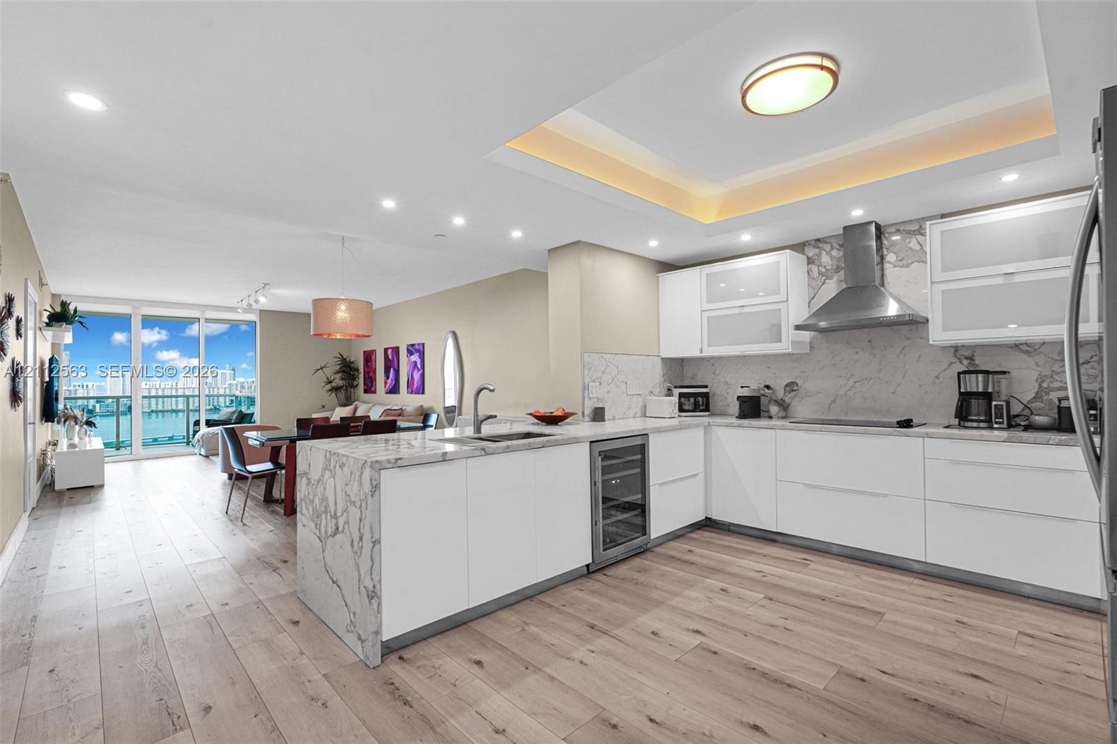 Kitchen featuring a waterfall-edge island, stone-finish countertops and backsplash, custom white cabinetry, built-in wine refrigeration, and recessed tray ceiling lighting