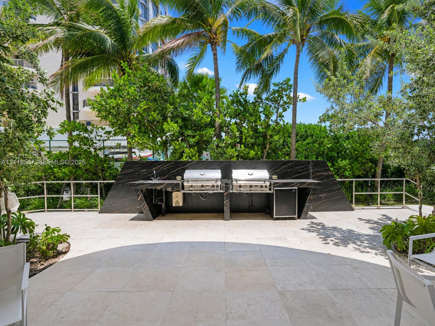Outdoor kitchen featuring a dark stone-finish backsplash and countertop, stainless steel dual grills, integrated refrigeration, and a stone-tiled patio surrounded by mature palm trees