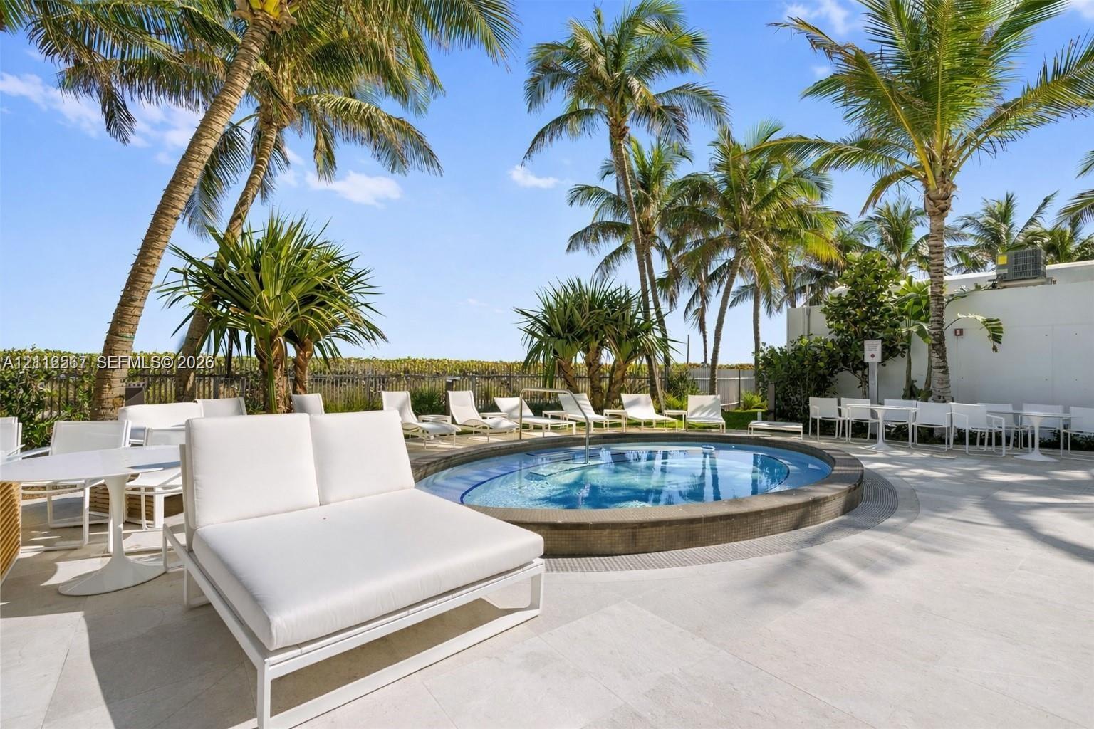 Circular in-ground spa featuring a stone-finish coping, surrounded by a paved deck, mature palm trees, and perimeter fencing