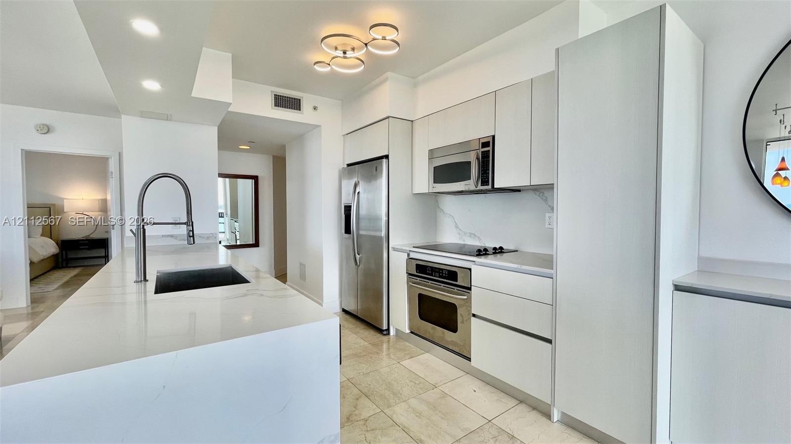 Kitchen featuring white flat-panel cabinetry, light-toned stone countertops, a waterfall-edge island, stainless steel appliances, and polished stone flooring