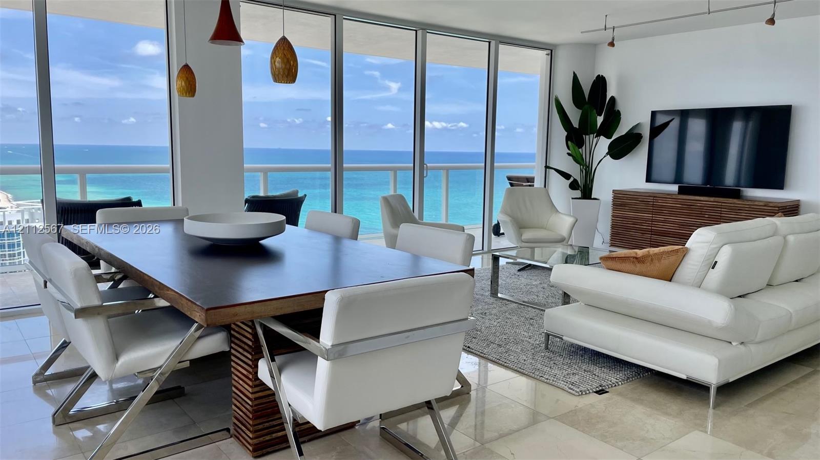 Open-concept living space featuring floor-to-ceiling glass walls, polished stone flooring, recessed track lighting, and a glass-railed balcony overlooking the ocean