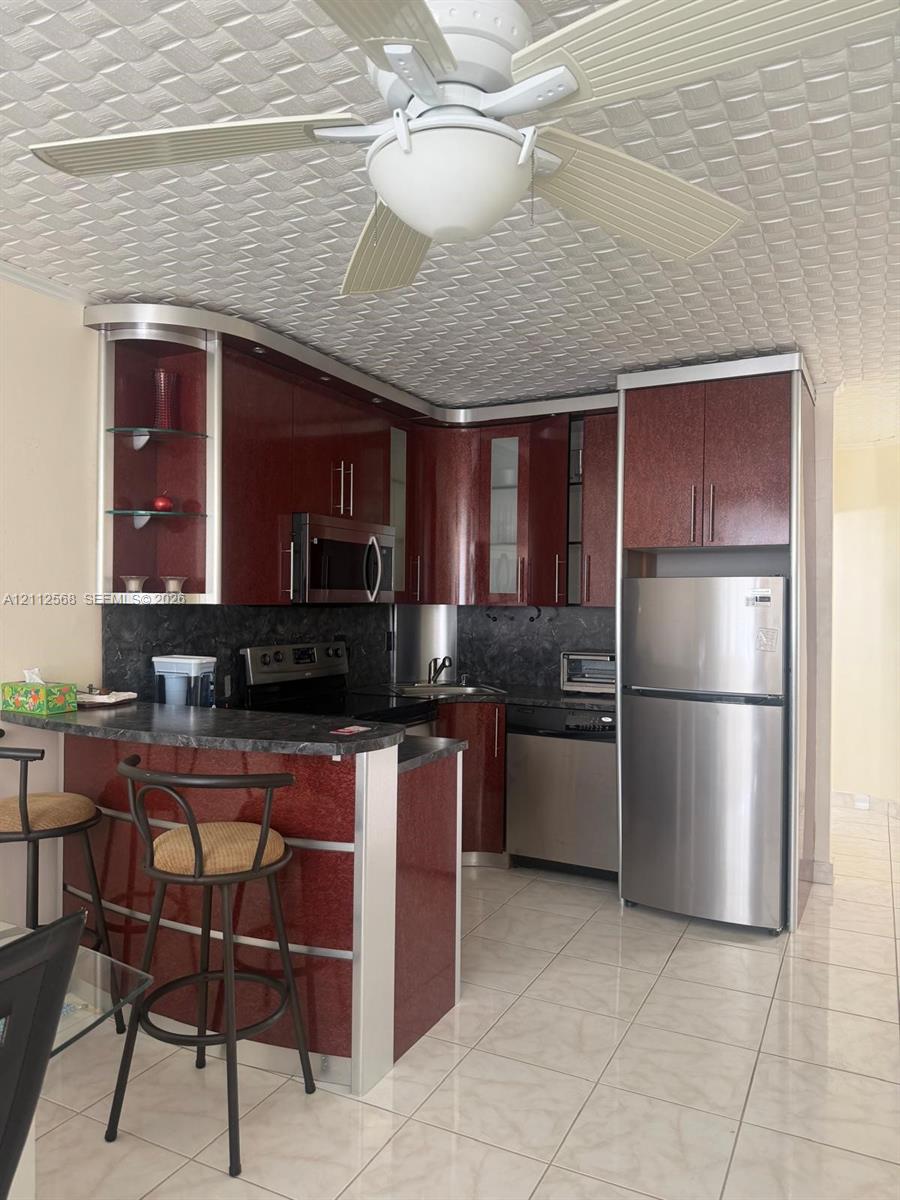 Kitchen featuring glossy cherry-toned cabinetry, stainless steel appliances, a peninsula counter with stone-finish surfaces, and a ceiling-mounted fan with integrated lighting