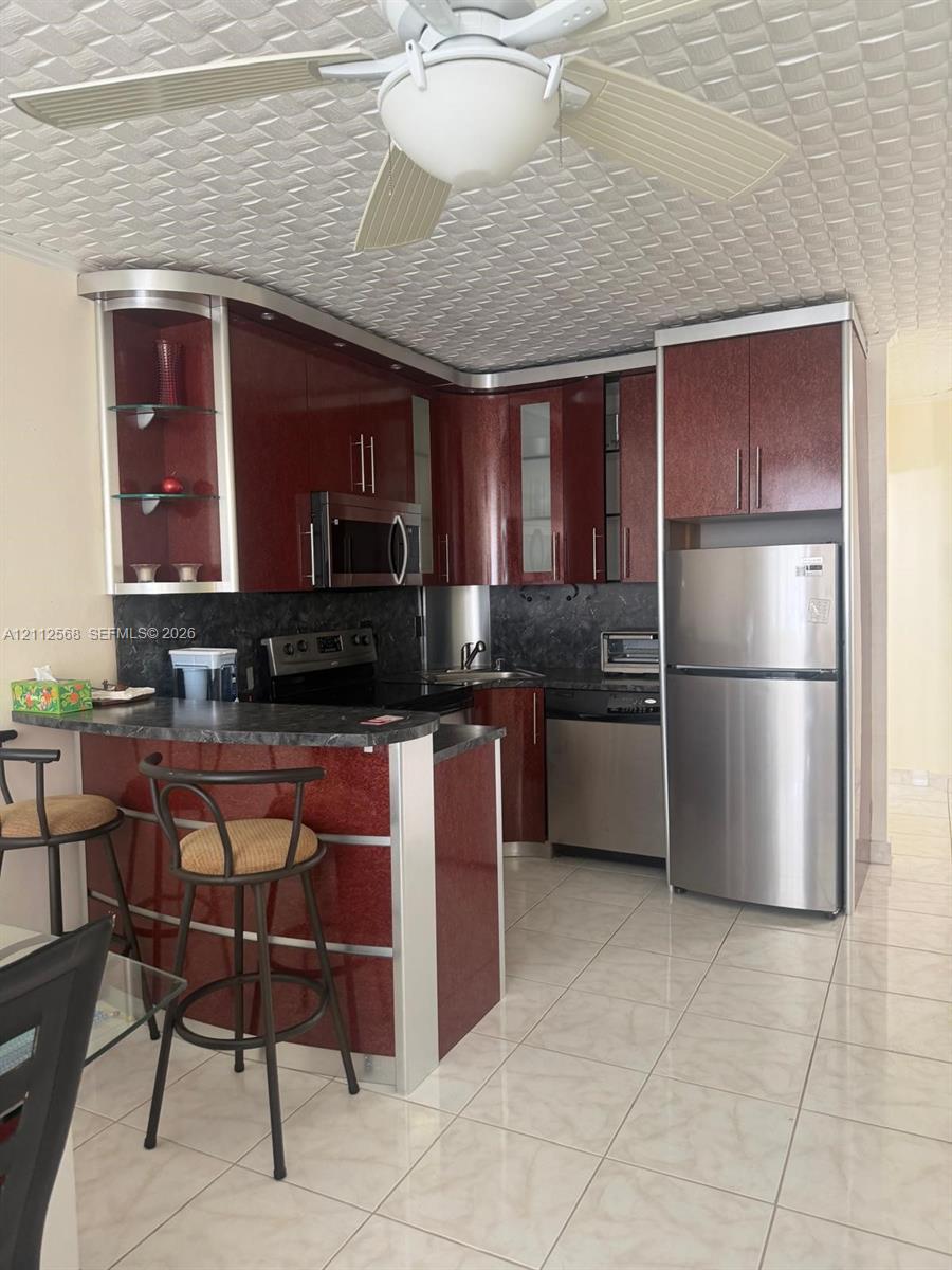 Kitchen featuring high-gloss cherry-finish cabinetry, stone-texture countertops, integrated stainless steel appliances, a peninsula breakfast bar, and tile flooring