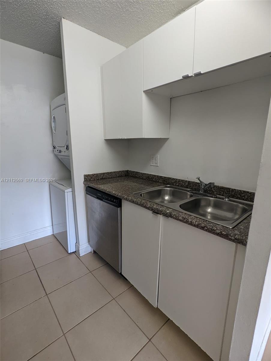 Utility space featuring white cabinetry, a dual-basin stainless steel sink, a stone-finish countertop, a stainless steel dishwasher, and stacked laundry appliances