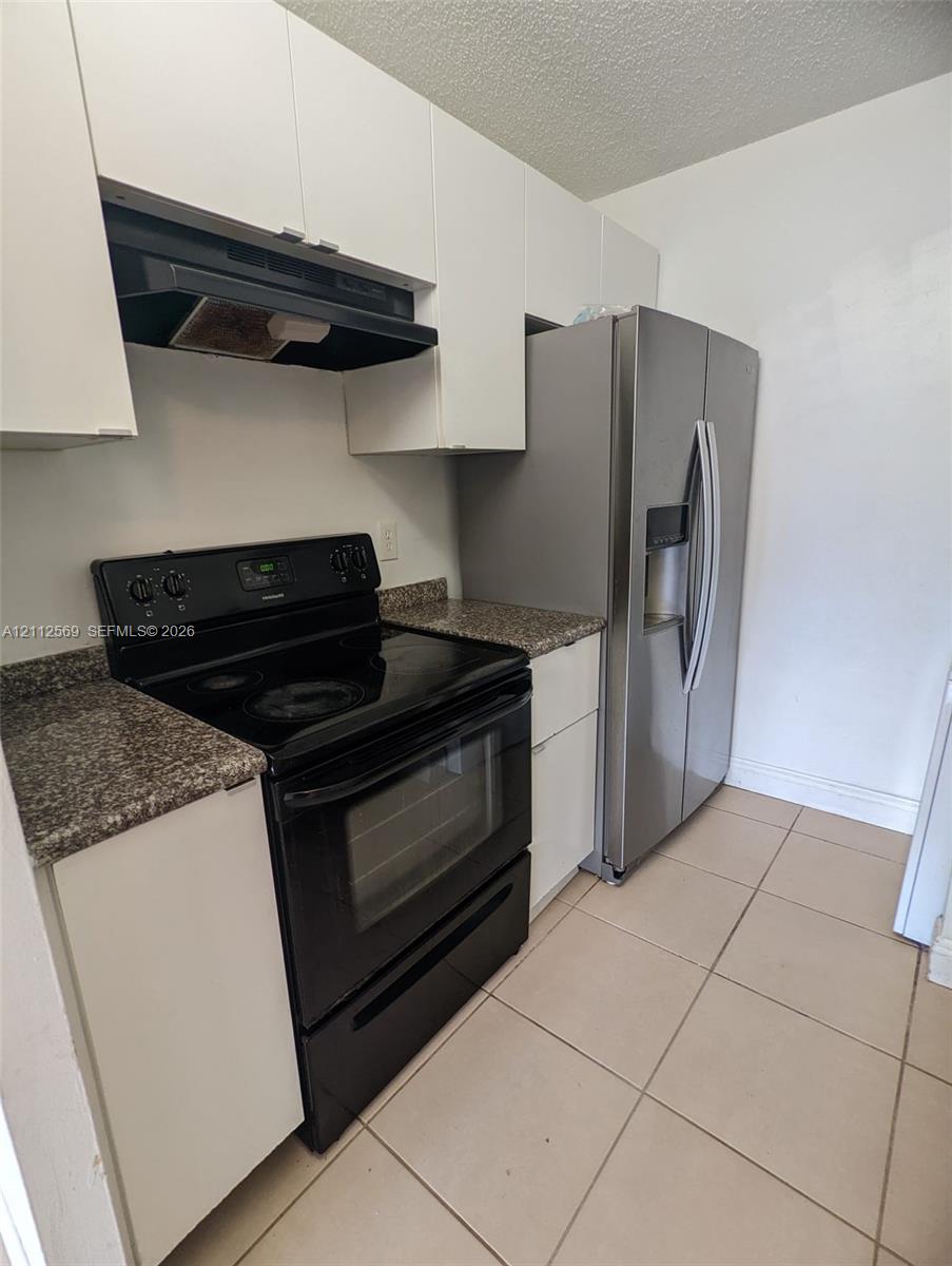 Kitchen featuring white cabinetry, stone-finish countertops, tile flooring, a stainless steel refrigerator, and a black electric range with a built-in exhaust hood