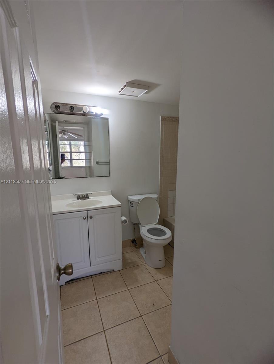 Vanity cabinet with integrated sink, rectangular wall-mounted mirror, multi-bulb light fixture, ceramic tile flooring, and standard toilet