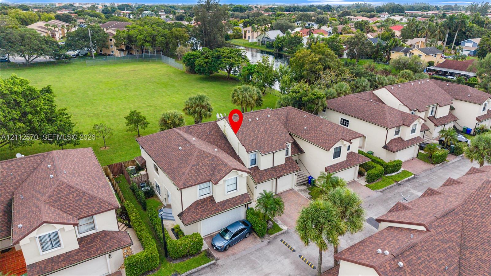 Aerial perspective of a multi-story residence featuring a shingled roof, stucco exterior, attached garage, and paved driveway, positioned amidst mature landscaping